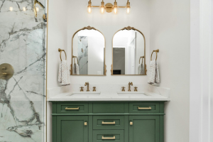 A modern bathroom with double vanity, green cabinets and gold overhead lighting.